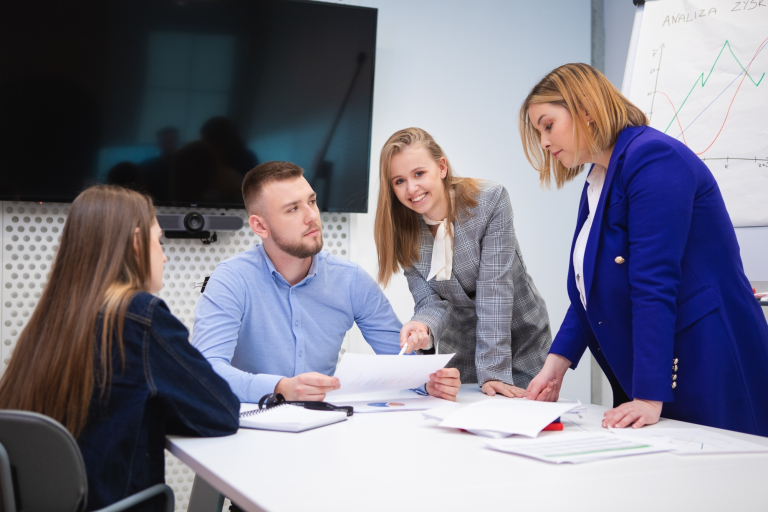 Psychologia zarządzania i biznesu | Studia jednolite magisterskie we Wrocławiu | Uniwersytet Dolnośląski DSW we Wrocławiu: Cztery osoby w sali – omawiają dokumenty i wykresy, jedna z kobiet pochylona nad stołem.