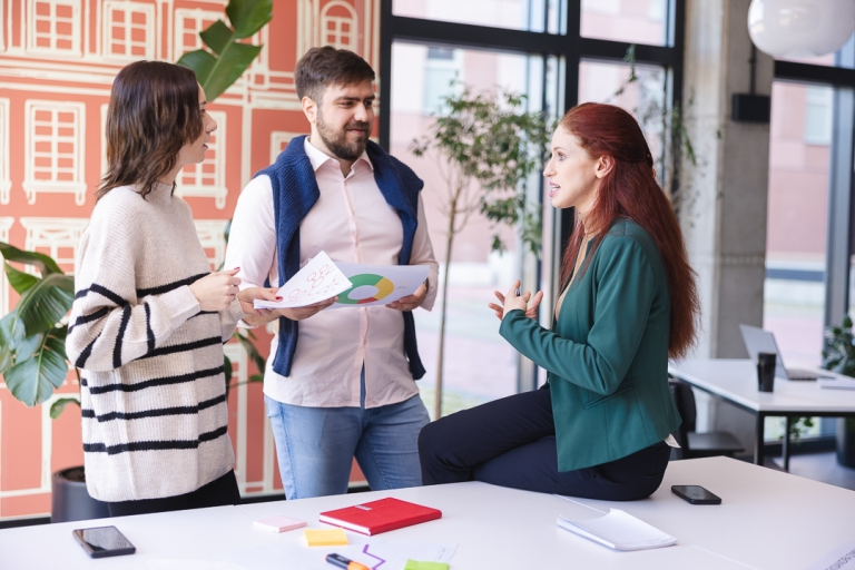 Growth & CRO Designer | Studia podyplomowe we Wrocławiu | Uniwersytet Dolnośląski DSW we Wrocławiu: Trzy osoby rozmawiają w jasnym, nowoczesnym biurze – kobieta siedzi na biurku, pozostali stoją z dokumentami w dłoniach.