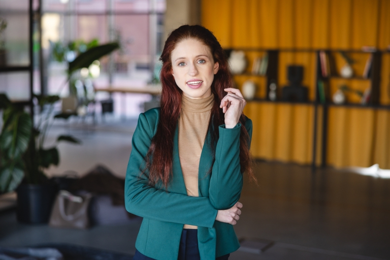 Centrum Wsparcia, Pomocy Psychologicznej i Psychoedukacji | Uniwersytet Dolnośląski DSW we Wrocławiu: Młoda kobieta z rudymi włosami uśmiecha się do obiektywu w przestrzeni coworkingowej.