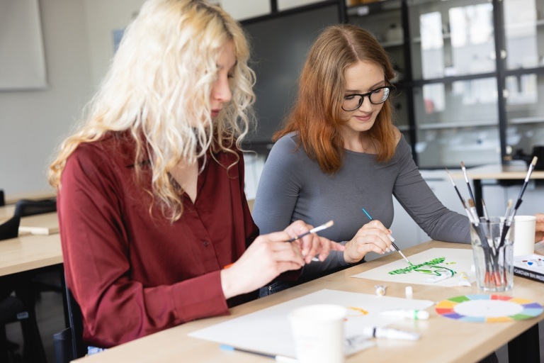 Terapia pedagogiczna | Studia I stopnia we Wrocławiu | Uniwersytet Dolnośląski DSW we Wrocławiu: Dwie studentki malujące przy stoliku w pracowni – jedna w bordowej koszuli, druga w szarej bluzce.