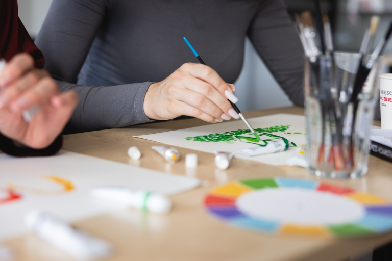 Terapia pedagogiczna | Studia I stopnia we Wrocławiu | Uniwersytet Dolnośląski DSW we Wrocławiu: Zbliżenie na dłonie osoby malującej zielony wzór farbą na papierze, w otoczeniu przyborów plastycznych.