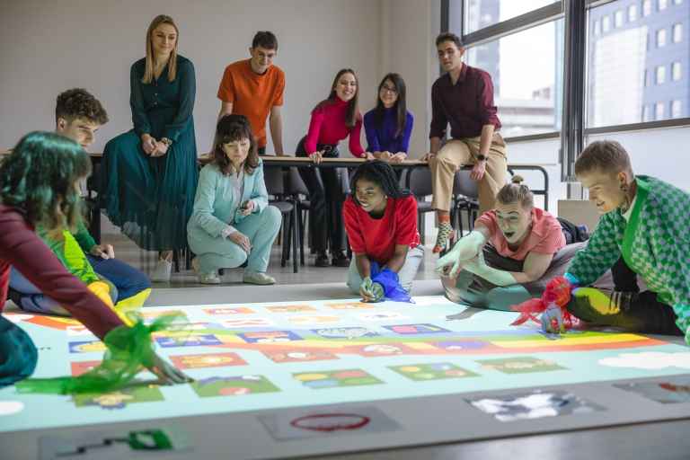 Pedagogika specjalna | Studia jednolite magisterskie we Wrocławiu | Uniwersytet Dolnośląski DSW we Wrocławiu:  Grupa studentów i wykładowców podczas interaktywnej zabawy edukacyjnej z wykorzystaniem kolorowej projekcji na podłodze.
