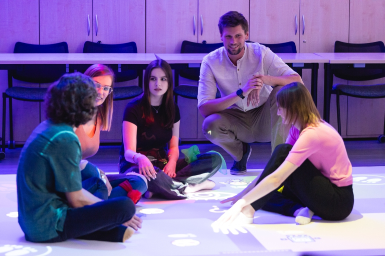 Pedagogika resocjalizacyjna | Studia I stopnia we Wrocławiu | Uniwersytet Dolnośląski DSW we Wrocławiu: Grupa młodych dorosłych siedzi na podłodze w sali edukacyjnej, uczestnicząc w zajęciach z wykorzystaniem interaktywnej podłogi sensorycznej.