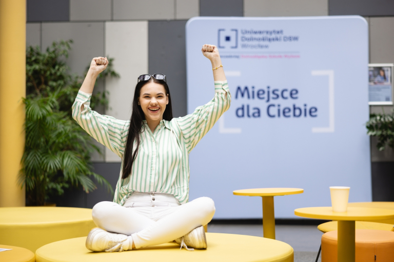 Komunikacja wizerunkowa | Studia II stopnia we Wrocławiu | Uniwersytet Dolnośląski DSW we Wrocławiu: Grafika główna: Uniwersytet Dolnośląski DSW, dla nowych studentów, studentka siedzi na sofie i trzyma ręce w górze w geście radości, w tyle napis na ścianie "Miejsce dla ciebie"