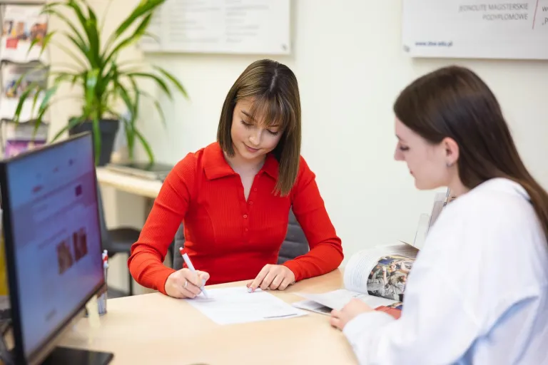 Stypendia i zniżki - studia I stopnia | Uniwersytet Dolnośląski DSW we Wrocławiu: Na zdjęciu dwie młode kobiety siedzą przy biurku w jasnym, nowoczesnym biurze. Kobieta w czerwonej bluzce podpisuje dokument, koncentrując się na tekście, podczas gdy druga kobieta, ubrana na biało, rozmawia z nią i trzyma otwartą broszurę. Na biurku znajdują się komputer, długopisy i inne dokumenty. W tle widać zieloną roślinę i tablicę informacyjną z napisem sugerującym, że to uczelniane biuro obsługi studentów.