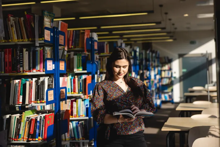Biblioteka | Uniwersytet Dolnośląski DSW we Wrocławiu: Na zdjęciu młoda kobieta stoi w bibliotece między regałami pełnymi książek. Ubrana w kolorową bluzkę w drobne kwiaty, czyta otwartą książkę, uśmiechając się lekko. Do wnętrza wpada naturalne światło, tworząc ciepłą i spokojną atmosferę sprzyjającą nauce. W tle widać puste stoliki i rzędy regałów.