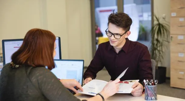 Biuro Karier i Praktyk | Uniwersytet Dolnośląski DSW we Wrocławiu: Młody mężczyzna w okularach siedzi przy biurku naprzeciwko kobiety, która pokazuje mu dokumenty. Na biurku stoją dwa monitory komputerowe i kubek z długopisami. W tle roślina i szafa z szufladami.