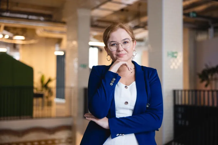 Zniżki i dofinansowania | Uniwersytet Dolnośląski DSW we Wrocławiu: Na zdjęciu kobieta w okularach, ubrana w granatową marynarkę i białą sukienkę, stoi w nowoczesnym, industrialnym wnętrzu. Uśmiecha się subtelnie, opierając dłoń na brodzie w zamyślonej pozie. W tle widoczne są elementy architektury z cegły, zielone akcenty roślin oraz oświetlenie sufitowe. Atmosfera profesjonalna i przyjazna.