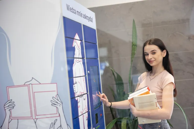 Biblioteka | Uniwersytet Dolnośląski DSW we Wrocławiu: Na zdjęciu młoda kobieta stoi przy maszynie do odbioru książek w bibliotece, oznaczonej napisem „Wiedza mnie uskrzydla!”. Ubrana w jasnoróżowy T-shirt i szare spodnie, uśmiecha się delikatnie, trzymając w ręku kilka książek. Drugą ręką obsługuje ekran dotykowy. W tle widoczna jest roślina doniczkowa i ściana z szarego kamienia. Atmosfera sprzyja nauce i korzystaniu z nowoczesnych udogodnień.