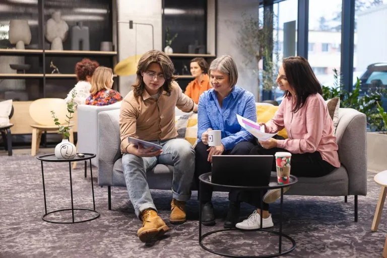 Dział Studiów Podyplomowych i Szkoleń | Uniwersytet Dolnośląski DSW we Wrocławiu: Na zdjęciu trzy osoby siedzą na szarej sofie w nowoczesnym, jasnym wnętrzu. Młody mężczyzna w okularach, beżowej koszuli i dżinsach pokazuje coś na tablecie dwóm kobietom. Kobieta w niebieskiej koszuli trzyma kubek, a druga, w różowej bluzce, przegląda dokumenty. Przed nimi stoi stolik z laptopem i kubkiem. W tle widać więcej osób rozmawiających oraz dużą witrynę z dekoracjami. Atmosfera sprzyja pracy zespołowej i wymianie pomysłów.