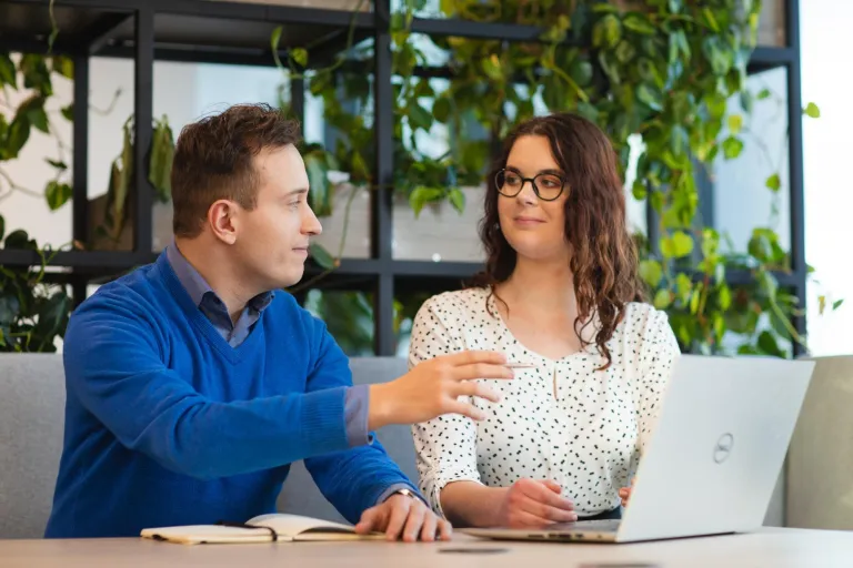 Studia II stopnia we Wrocławiu | Uniwersytet Dolnośląski DSW we Wrocławiu: Mężczyzna w niebieskim swetrze i kobieta w okularach pracują razem przy stole z laptopem w nowoczesnym wnętrzu z roślinami w tle. Oboje rozmawiają i analizują notatki.
