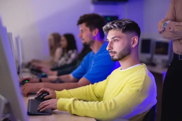 Media kreatywne – projektowanie gier i animacji | Studia II stopnia we Wrocławiu | Uniwersytet Dolnośląski DSW we Wrocławiu: Główna grafika: Inżynier aplikacji i systemów chmurowych  - student w żółtym swetrze robiący zadanie przy komputerze