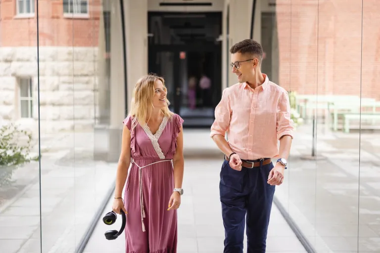 Pedagogika | Studia I stopnia we Wrocławiu | Uniwersytet Dolnośląski DSW we Wrocławiu: Na zdjęciu kobieta i mężczyzna idą korytarzem w przeszklonym budynku. Oboje uśmiechają się do siebie i prowadzą rozmowę. Kobieta w różowej sukience trzyma w ręce słuchawki, mężczyzna ma na sobie jasnoróżową koszulę i granatowe spodnie. W tle widoczne są ceglane i kamienne elewacje budynków oraz ławki, co tworzy przyjazną atmosferę kampusu.