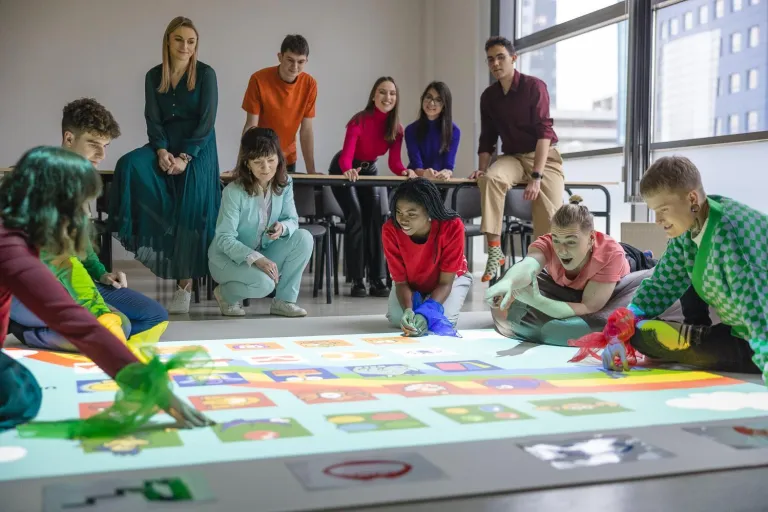 Pedagogika | Studia I stopnia we Wrocławiu | Uniwersytet Dolnośląski DSW we Wrocławiu: Grupa studentów i wykładowców bierze udział w zajęciach z wykorzystaniem maty interaktywnej. Część osób klęczy lub siedzi na podłodze, wskazując elementy wyświetlane na macie, inni obserwują z uśmiechami w tle. Atmosfera sprzyja zaangażowanej nauce i zabawie.