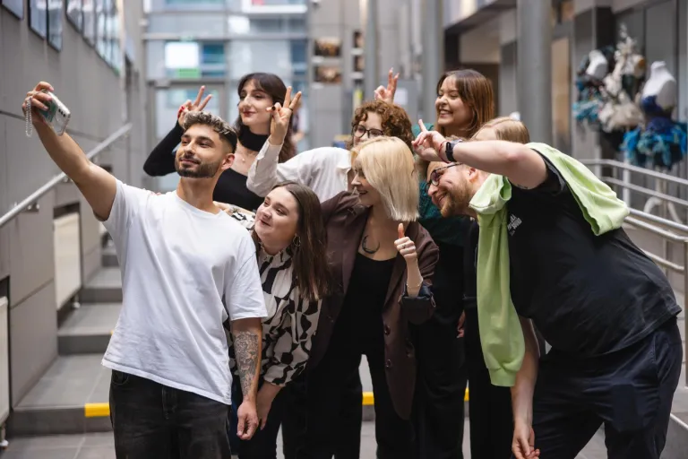 Zapraszamy na sesję zdjęciową | 25-26.02.2026 | Aktualności | Uniwersytet Dolnośląski DSW we Wrocławiu: Grupa młodych osób pozuje do wspólnego selfie w jasnym korytarzu uczelni. Wszyscy uśmiechają się, robią gesty pokoju i radośnie spoglądają w stronę telefonu trzymanego przez mężczyznę z przodu.