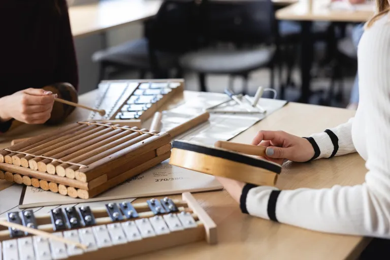 Pedagogika opiekuńczo-wychowawcza z elementami socjoterapii | Studia I stopnia we Wrocławiu | Uniwersytet Dolnośląski DSW we Wrocławiu: Zbliżenie na dłonie dwóch osób grających na instrumentach perkusyjnych – bębenku, dzwonkach i instrumentach drewnianych. Na stole leżą nuty i pałeczki, co wskazuje na zajęcia z zakresu muzykoterapii lub edukacji muzycznej.