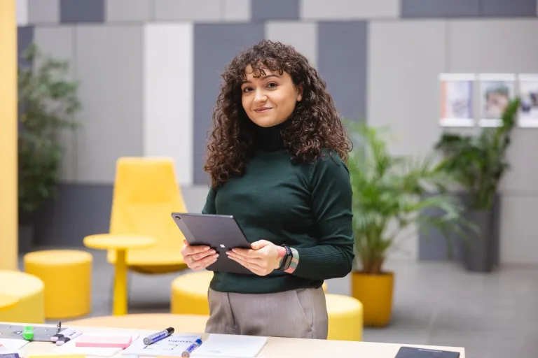 Marketing i social media | Studia I stopnia we Wrocławiu | Uniwersytet Dolnośląski DSW we Wrocławiu: Kobieta z kręconymi włosami w zielonym golfie trzyma tablet i uśmiecha się lekko. Za nią kolorowe, nowoczesne wnętrze z żółtymi meblami i roślinami. Na stole widać dokumenty i przybory do pisania.