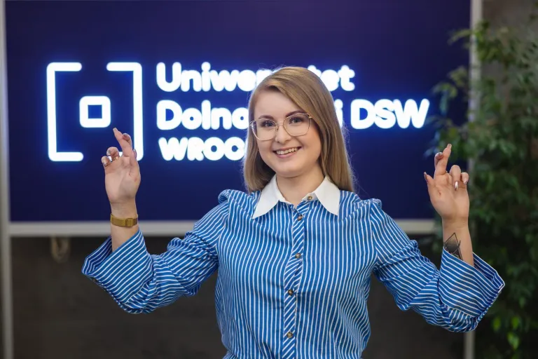 Zarządzanie nieruchomościami | Studia I stopnia we Wrocławiu | Uniwersytet Dolnośląski DSW we Wrocławiu: Młoda kobieta w niebieskiej koszuli w paski stoi przed logotypem Uniwersytetu Dolnośląskiego DSW we Wrocławiu. Uśmiecha się i pokazuje gest skrzyżowanych palców, trzymając kciuki za powodzenie.