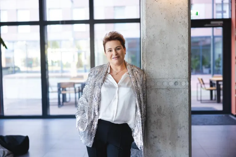 Marketing i social media | Studia I stopnia we Wrocławiu | Uniwersytet Dolnośląski DSW we Wrocławiu: „Kobieta w błyszczącej marynarce i białej koszuli opiera się o betonowy filar w nowoczesnym wnętrzu z dużymi oknami.”