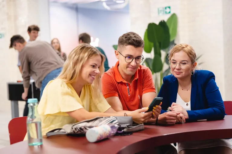 Zarządzanie | Studia I stopnia we Wrocławiu | Uniwersytet Dolnośląski DSW we Wrocławiu: „Trzy uśmiechnięte osoby siedzą przy stole i wspólnie przeglądają treści na telefonie w przestrzeni coworkingowej.”