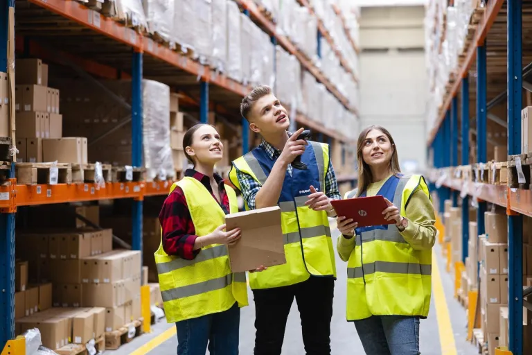 Logistyka | Studia I stopnia we Wrocławiu | Uniwersytet Dolnośląski DSW we Wrocławiu: Studenci logistyki w neonowych kamizelkach spoglądają wspólnie na półki z kartonami