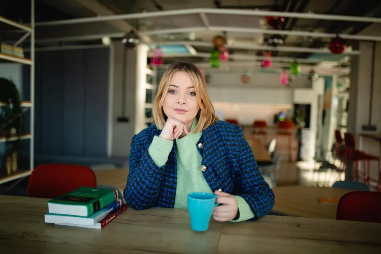 Wyniki wyszukiwania | Uniwersytet Dolnośląski DSW we Wrocławiu: Kobieta w niebieskim żakiecie siedzi przy stole z kubkiem w dłoni. Przed nią leżą książki i notatki. W tle kolorowe dekoracje i regały z książkami.