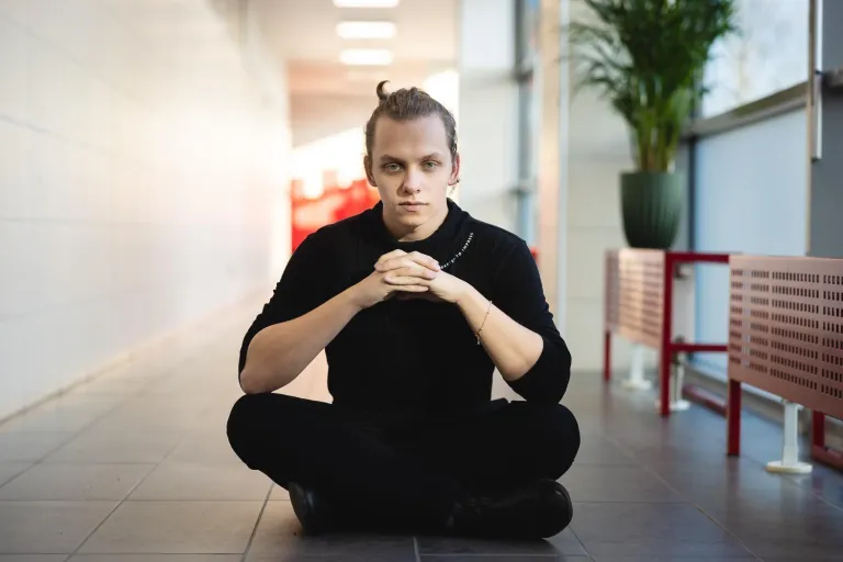 Młody mężczyzna w czarnym stroju siedzi po turecku na podłodze jasnego korytarza uczelni, patrząc prosto w obiektyw z złożonymi rękami, wyrażając pewność siebie i skupienie.