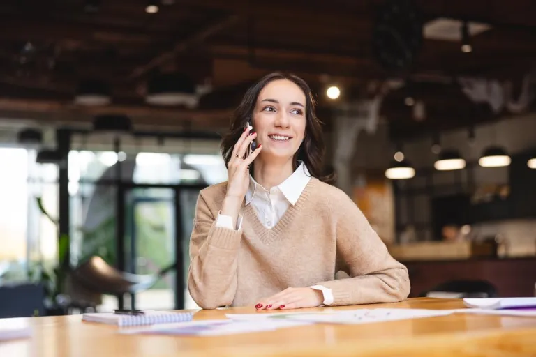 HR Business Partner z elementami coachingu | Studia I stopnia we Wrocławiu | Uniwersytet Dolnośląski DSW we Wrocławiu: Kobieta w beżowym swetrze rozmawia przez telefon, siedząc przy stole w nowoczesnym biurze lub kawiarni. Uśmiecha się, a przed nią leżą notatki i dokumenty.