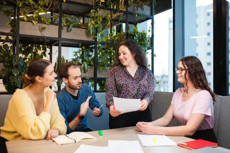 Psychologia sądowa | Studia jednolite magisterskie we Wrocławiu | Uniwersytet Dolnośląski DSW we Wrocławiu: Czworo studentów przy stole – kobieta w kolorowej bluzce trzyma kartkę i uśmiecha się do reszty.
