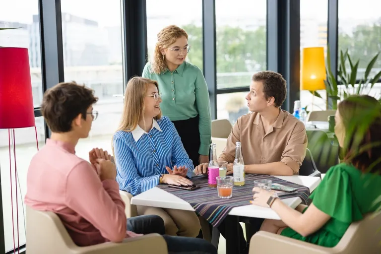 Pedagogika resocjalizacyjna | Studia I stopnia we Wrocławiu | Uniwersytet Dolnośląski DSW we Wrocławiu: Grupa pięciu studentów siedzi przy stole w jasnej, nowoczesnej przestrzeni. Rozmawiają i uśmiechają się, na stole stoją butelki z napojami i szklanki.