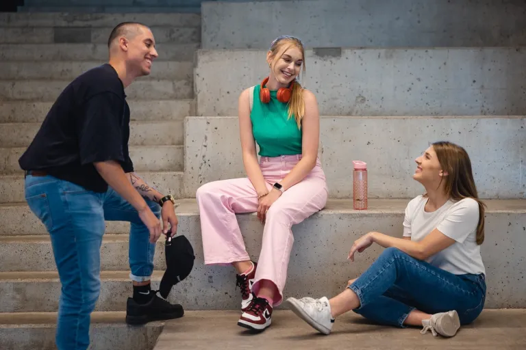 Wyniki wyszukiwania | Uniwersytet Dolnośląski DSW we Wrocławiu: Troje młodych ludzi siedzi na betonowych schodach, uśmiecha się i rozmawia w swobodnej atmosferze.