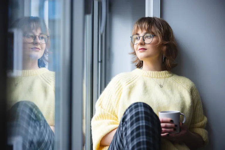 Szkoły partnerskie | Uniwersytet Dolnośląski DSW we Wrocławiu: Studentka siedzi na parapecie i patrzy za okno trzymając filiżankę napoju w ręce